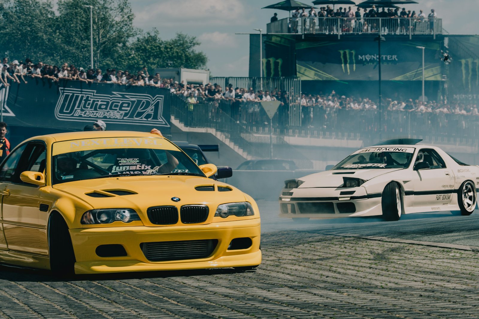 A yellow BMW and a white Mazda RX-7 drifting side by side through a corner, tyre smoke pouring off the rear wheels