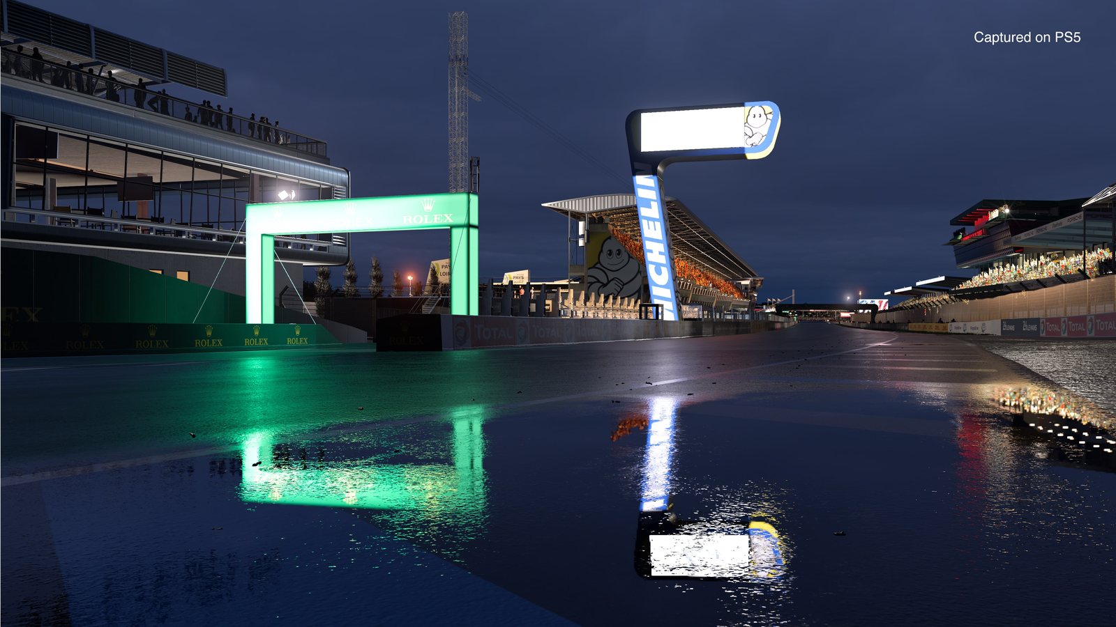 A pit lane at night under neon light, wet tarmac reflecting the gantry